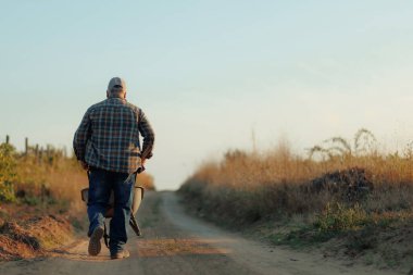 Odaklanmış bir ifadeyle kıdemli bir çiftçi, toprak bir yolda el arabasını itiyor. Çevresindeki alan canlı ve yeşil, çiftçiliğin sıkı çalışmasını vurguluyor..