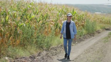 Güneşli bir mısır tarlasının yanında bir kadın çiftçi. Altın mısır sapları ve mavi gökyüzü kırsal yaşamın huzurlu ve huzurlu bir görüntüsünü yaratır..