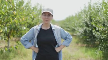 Bir meyve bahçesinde, kafasında şapka olan genç bir dişi çiftçi gelişmekte olan bitkileri inceler.