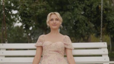 Uzun saçlı neşeli bir sarışın kadın, gülümsüyor ve salıncakta rahatlıyor, sıcak güneş ışığı güzelliğini arttırıyor..