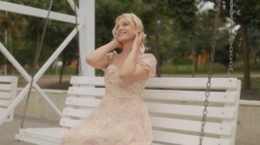 Sarışın, orta yaşlı bir kadın salıncakta oturuyor, gün ışığında sıcak bir şekilde gülümsüyor, kaygısız ifadesi anın keyfini çıkarıyor..