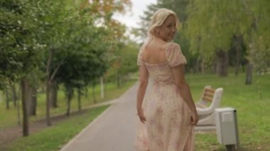 Pembe elbiseli genç sarışın bir kadın park caddesinde ağır çekimde yürüyor, kameraya neşeyle gülümsüyor, mutluluk ve güven saçıyor..