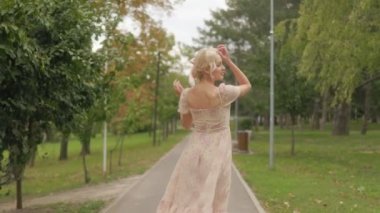 Pembe elbiseli neşeli sarışın bir kadın, bir park yolunda zarifçe dönüyor. Parlak gülüşü kameraya doğru dönerken parlıyor..