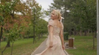 Genç sarışın bir kadın güvenli bir şekilde kameraya gülümsüyor. Parkta yürürken, güneşli gökyüzünün altında pembe elbisesiyle mutlu bir şekilde dönüyor..