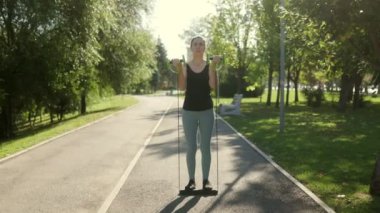 Genç esmer bir kadın güneşli bir günde parkta direnç bantları kullanarak spor yapıyor. Açık hava egzersizlerini, canlılığı ve kendine güveni sever..