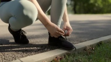 Kadın, spor salonunu bitirdikten sonra ayakkabı bağcıklarını bağlıyor. Parkta güneş ışığı altında, aktif bir yaşam tarzı sürdürüyor..