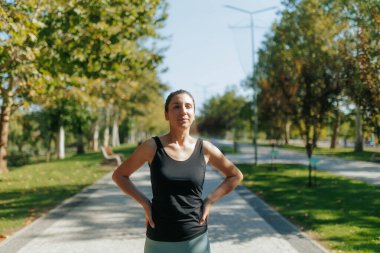 Sportif esmer bir kadın, spor yaptıktan sonra parkta dinleniyor. Parlak güneşin altında yakalanmış. Rahatlamış ifadesi gençlik güvenini ve enerjisini vurguluyor..