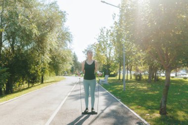 Formda genç bir kadın parkta direnç bandı egzersizleri yapıyor. Açık havanın ve güneşli günün sıcaklığının tadını çıkarırken spor amaçlarına odaklanıyor..