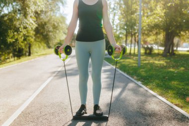 Formda genç bir kadın parkta direnç bandı egzersizleri yapıyor. Açık havanın ve güneşli günün sıcaklığının tadını çıkarırken spor amaçlarına odaklanıyor..