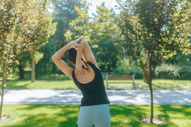 Güneşli bir parkta genç bir kadının fiziksel fitness rutinine bakışı. Atletik kıyafetler giyiyor ve duruşu güven ve gücü yansıtıyor.