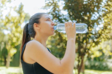 Genç beyaz bir kadın güneşli bir günde parkta spor yaptıktan sonra şişeden su içer ve fiziksel egzersizden sonra susuz kalmaz..