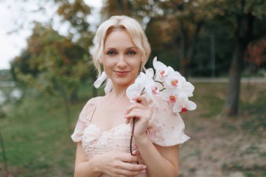 Elinde bir orkide çiçeği tutan sarışın bir kadının portresi. Açık hava ortamında kameraya sıcak bir şekilde gülümsüyor. Onun ifadesi neşeli ve flörtöz, zarafet ve pozitiflik saçıyor.