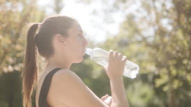 Genç beyaz bir kadın güneşli bir günde parkta spor yaptıktan sonra şişeden su içer ve fiziksel egzersizden sonra susuz kalmaz..
