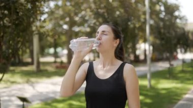 Güneşli bir parkta jimnastik eğitiminden sonra su şişesinden su içen genç bir kadın temiz havanın tadını çıkarıyor..
