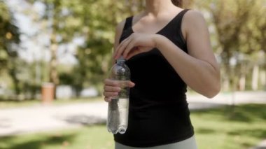 Spor yaptıktan sonra elinde bir şişe su tutan bir kadın, güneşli bir günde açık hava eğitiminin spor ve aktif yaşam tarzını yakalıyor..