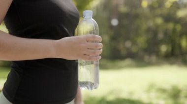 Açık hava eğitimi sırasında canlılık, zindelik ve susuzluğu temsil eden jimnastik egzersizlerinden sonra su şişesini tutan genç bir kadının yakın plan çekimi..