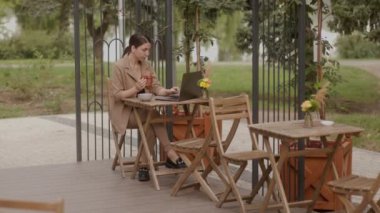 Bir iş kadını terasta uzaktan çalışıyor, laptopunda düşünceli bir ifadeyle yazıyor, elinde bir kruvasan tutuyor ve kahvaltıda çay içiyor..