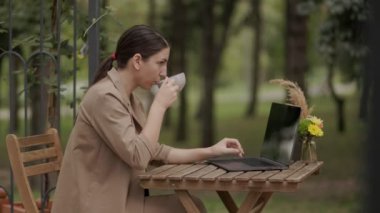 Genç ve ciddi esmer bir kadın terasta bir masada oturuyor, elinde bir fincan çay tutarken dizüstü bilgisayarda yazıyor. İşine odaklanmış, kahvaltısını hazırlıyor.