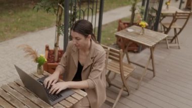 Takım elbise giymiş genç bir esmer girişimci bilgisayarında uzaktan çalışıyor, teras masasında oturuyor, mesleki görevlerine dalmış..