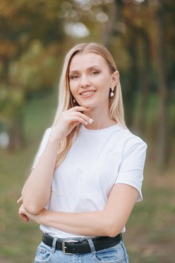 Huzurlu bir park ortamında, orta yaşlı, mutlu, sarışın bir kadın doğrudan kameraya bakar.,