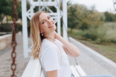 Sarışın, orta yaşlı bir kadın parkta salıncakta sallanıyor. Bakışları kameraya çevrilmiş. Parlak gülümsemesi rahat ve kaygısız havasını yansıtıyor..