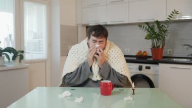 Genç beyaz bir adam evde griple savaşıyor, battaniyeye sarılmış ve mutfak sandalyesine oturmuş hapşırıyor. 