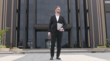 Siyah bir duvara karşı sakallı ve gözlüklü, takım elbiseli genç bir iş adamı. Dizüstü bilgisayarla duruyor.