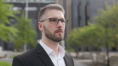 Siyah bir duvara karşı sakallı ve gözlüklü, takım elbiseli genç bir iş adamı. Etrafa bakıyor. 