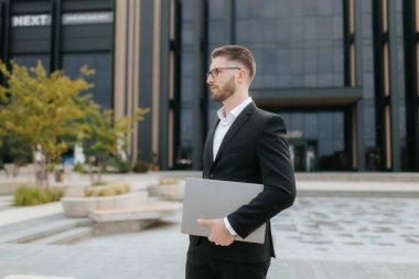 Gözlüklü ve sakallı genç bir şirket çalışanı bir ofis binasının önünde dizüstü bilgisayar tutuyor. Onun ciddi ifadesi kararlılığı ve odaklanmayı yansıtır..