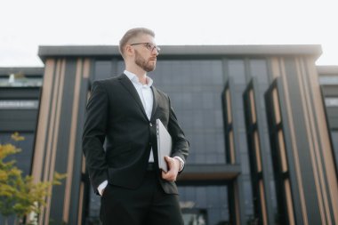 Sakallı, gözlüklü ve şık takım elbiseli profesyonel bir adam iş yerindeki bir ofis binasının önünde dizüstü bilgisayarıyla duruyor. Ciddi ve azimli bir tavır sergiliyor..