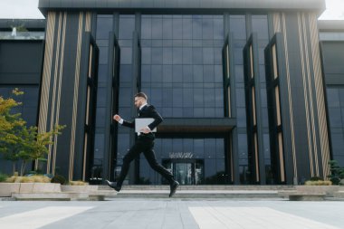 Ciddi bir genç iş adamı, sakallı ve gözlüklü bir şirket binasının önünde dizüstü bilgisayarını tutarak koşuyor..