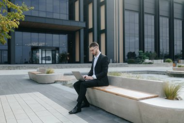 Şık takım elbiseli genç bir işadamı, modern bir ofis binasının önünde dizüstü bilgisayarıyla çalışıyor. Şehir merkezindeki kalabalık bir iş bölgesinde güven ve verimlilik yayıyor..
