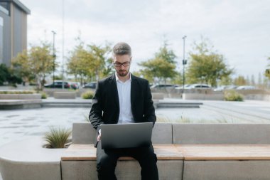 Young white-collar worker in a formal suit sits outdoors, working on his laptop in front of a modern office building, representing efficiency in a fast-paced urban environment. clipart