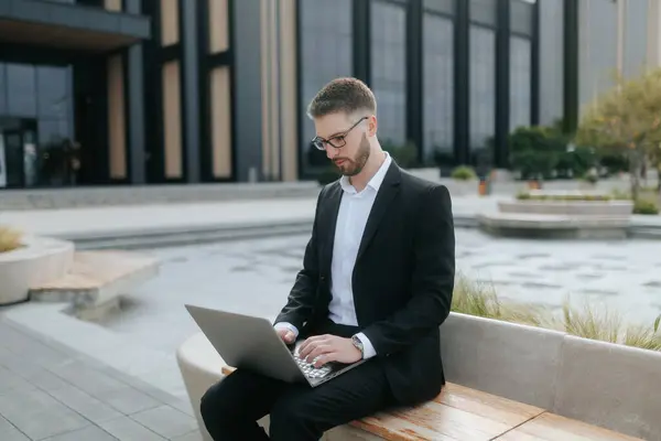 stock image Young businessman in a sleek suit working on his laptop outside a modern office building. He is focused, showcasing productivity and confidence in a contemporary urban setting.