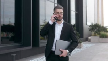 Siyah bir duvara karşı sakallı ve gözlüklü, takım elbiseli genç bir iş adamı. Akıllı telefondan konuşuyor. 