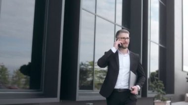 Siyah bir duvara karşı sakallı ve gözlüklü, takım elbiseli genç bir iş adamı. Akıllı telefondan konuşuyor. 