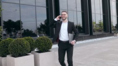 Siyah bir duvara karşı sakallı ve gözlüklü, takım elbiseli genç bir iş adamı. Akıllı telefondan konuşuyor ve yürüyor. 