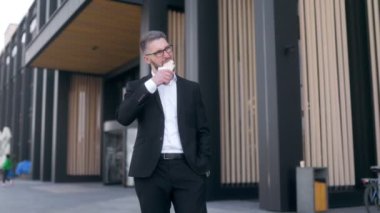 Siyah bir duvara karşı sakallı ve gözlüklü, takım elbiseli genç bir iş adamı. Sandviç yiyor. 