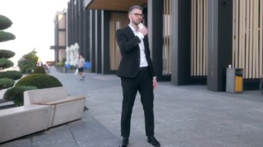 Siyah bir duvara karşı sakallı ve gözlüklü, takım elbiseli genç bir iş adamı. Sandviç yiyor. 