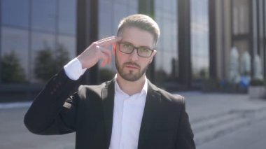 Siyah bir duvara karşı sakallı ve gözlüklü, takım elbiseli genç bir iş adamı. Kameraya dikkatle bakıyor, silah işareti yapıyor.