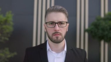 Siyah bir duvara karşı sakallı ve gözlüklü, takım elbiseli genç bir iş adamı. Düşünceli bir şekilde kameraya bakıyor, başını sallıyor. 