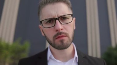 Siyah bir duvara karşı sakallı ve gözlüklü, takım elbiseli genç bir iş adamı. Düşünceli bir şekilde kameraya bakıyor.