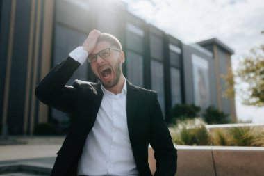 Takım elbiseli ve gözlüklü genç bir profesyonel bir ofis binasının önünde duruyor. Telefonundan kötü haber aldıktan sonra öfkeden ve yorgunluktan bağırıyor..