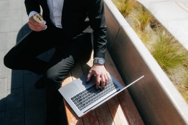 Takım elbiseli bir adam önündeki dizüstü bilgisayarla bankta oturuyor. Dizüstü bilgisayarı üzerinde çalışırken bir sandviç yiyor.