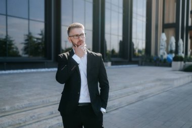 Sakallı ve gözlüklü bir işadamı, şık bir takım elbise içinde, ofis kulesinin önünde duruyor, odaklanmış bir ifadeyle önemli mesleki kararları düşünüyor..