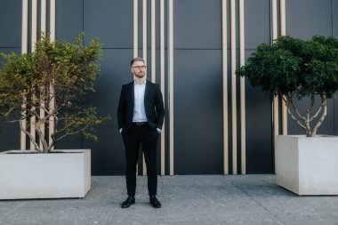 İş elbisesi ve gözlüklü sakallı profesyonel bir adam karanlık bir duvara karşı ciddi bir ifadeyle poz veriyor. İş dünyasında başarı ve prestiji temsil ediyor..