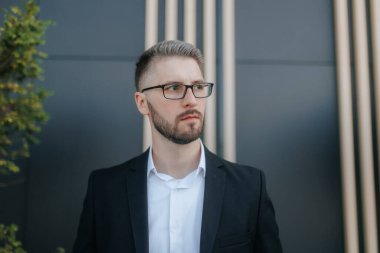 Şık bir takım elbise ve gözlük takmış kendine güvenen bir iş lideri, karanlık bir zemine karşı duruyor. Sakallı yüzü ve ciddi ifadesi kurumsal otoritenin özünü yansıtıyor..