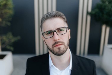 Ciddi ve şüpheli bir adam, sakallı ve gözlüklü, resmi bir takım elbise giymiş, karanlık bir duvarın karşısında duruyor. Düşünceli bir şekilde kameraya bakıyor, profesyonel bir davranışı yansıtıyor..