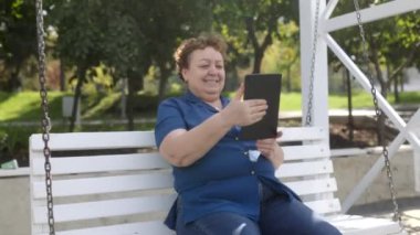 Parkta salıncakta sallanan tablet bilgisayar kullanan yaşlı bir kadının portresi 
