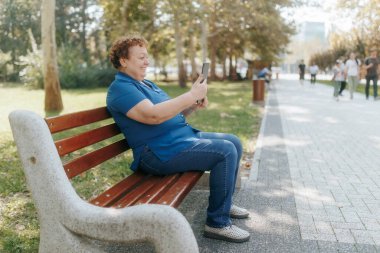 Yaşlı bir kadın parktaki bir bankta rahatça oturur tabletine bağlanırken sevinçle gülümser ve çevreleyen doğanın güzelliğinin ve dijital etkileşimin tadını çıkarır..